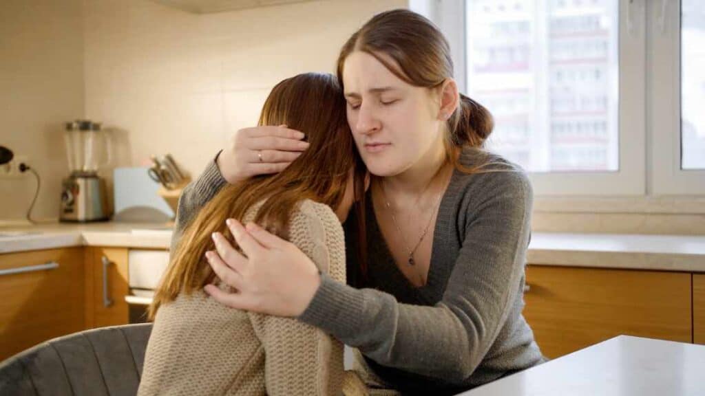 Supportive woman hugs distressed teen in kitchen, showing empathy, key to motivating teens with depression through care and connection.