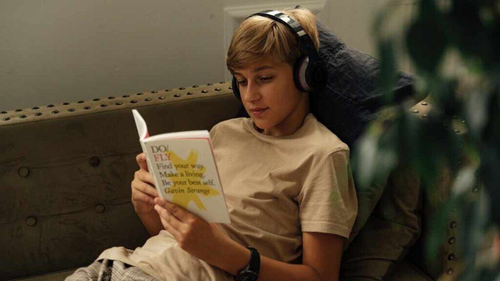 Teen boy in residential treatment relaxes on couch, wearing headphones and reading a motivational book, showing personal growth.
