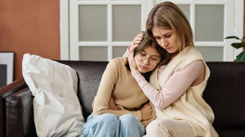 Worried mother hugs sad teen on couch, capturing the fine line between supportive parenting and unintentional pressure.